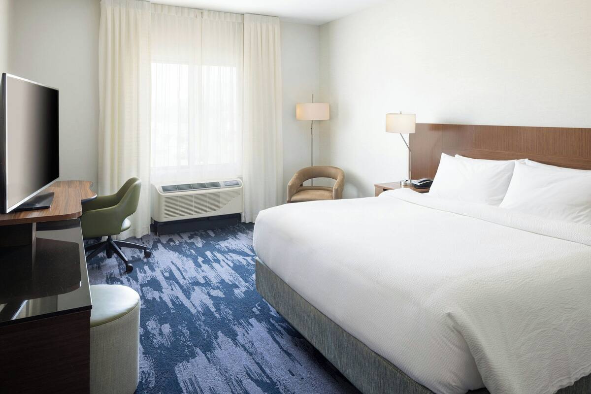 A hotel room with a large bed, a desk with a flat-screen TV, a chair, a lamp, and a window with curtains.
