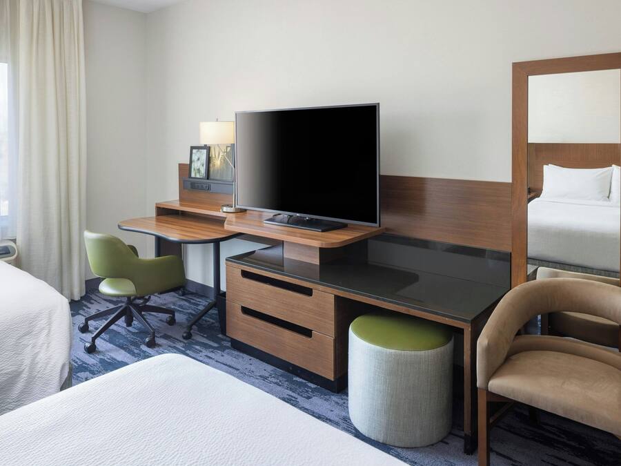 A hotel room with a bed, a desk with a flat-screen TV, a green office chair, a wooden cabinet, and a large mirror.