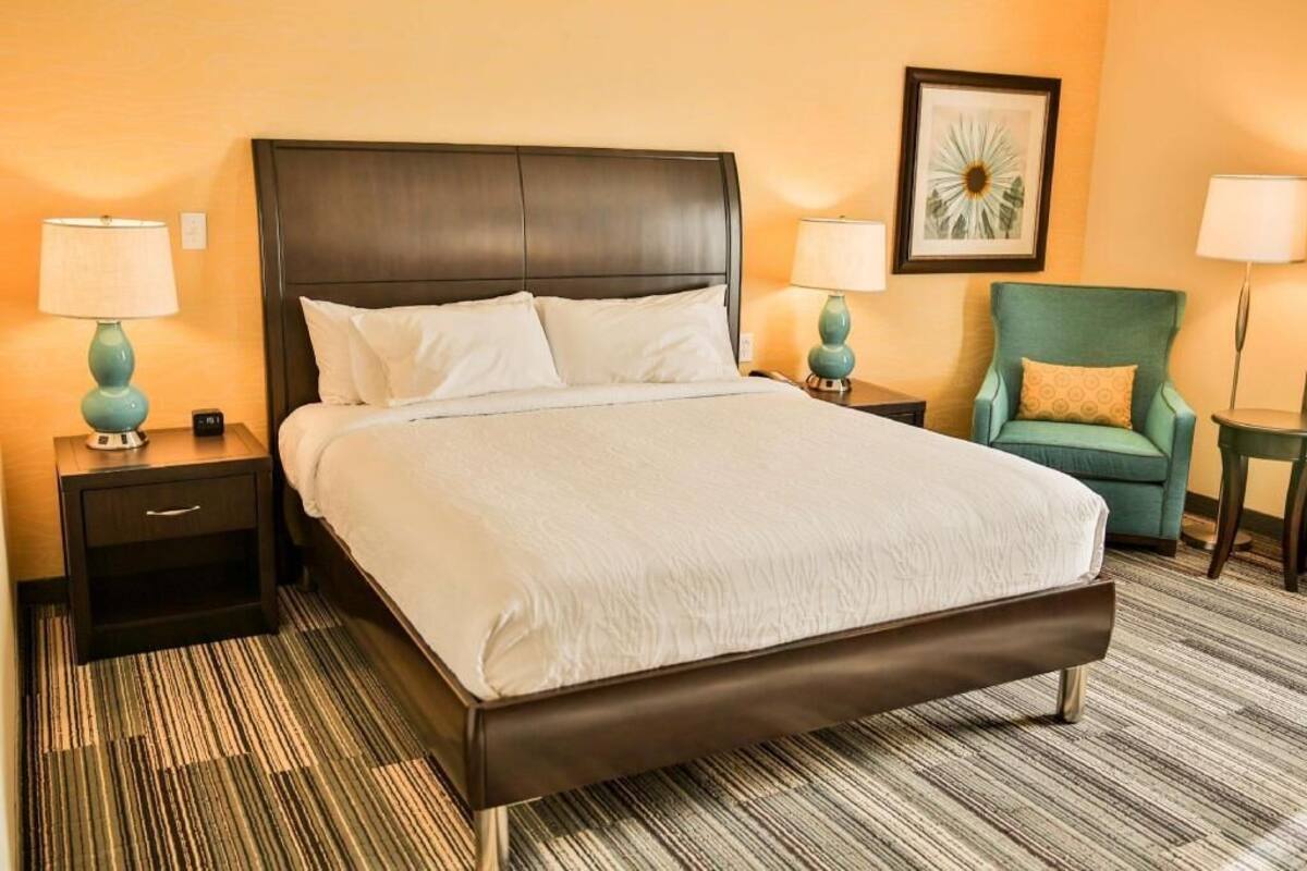 A hotel room with a large bed, two bedside tables with lamps, a green armchair, and a framed picture on the wall.