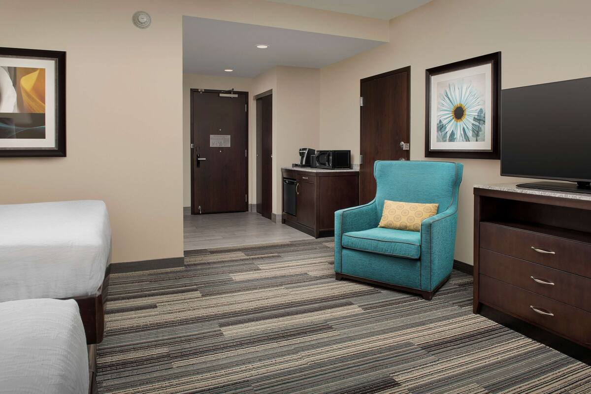 A hotel room with a bed, a teal armchair, a flat-screen TV, a coffee maker, and a framed artwork on the wall.