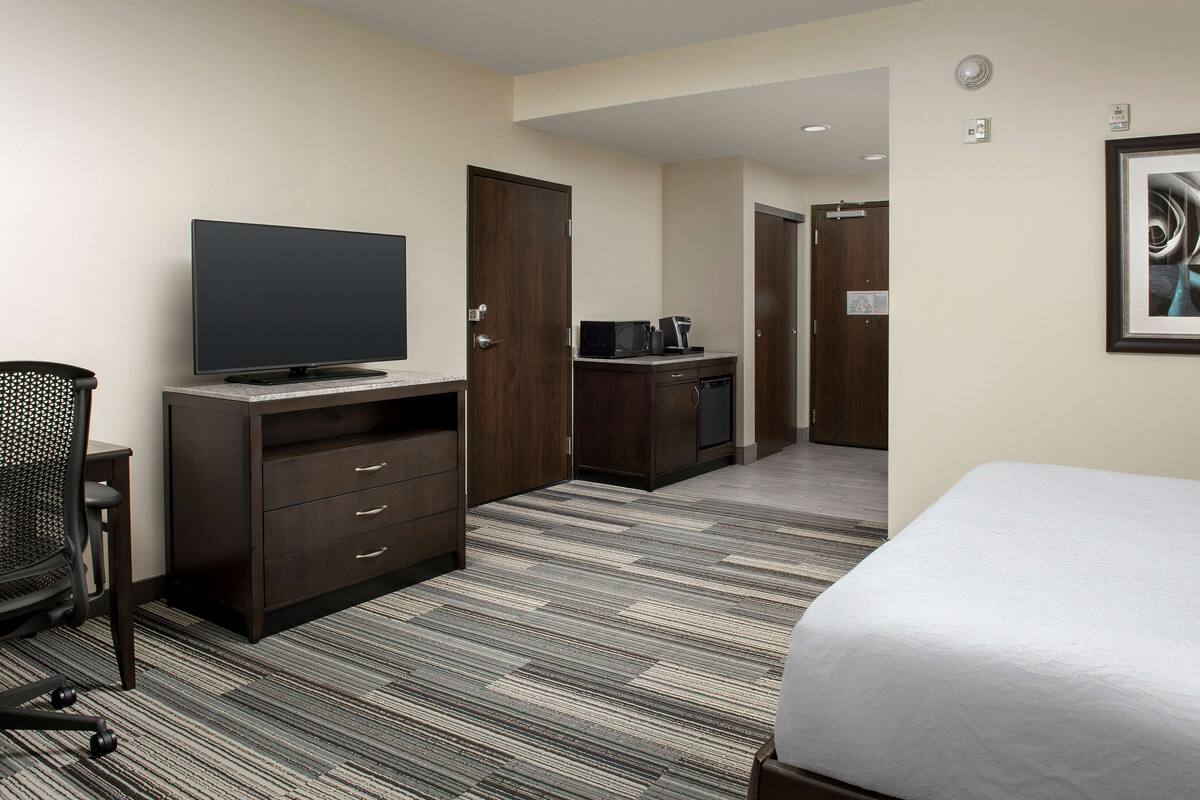A hotel room with a bed, a TV, a desk with a coffee maker, and a chair.