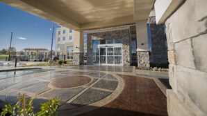 A modern building entrance with a stone facade, glass doors, and a paved area with a circular design. - Hilton Garden Inn Murfreesboro (Murfreesboro)