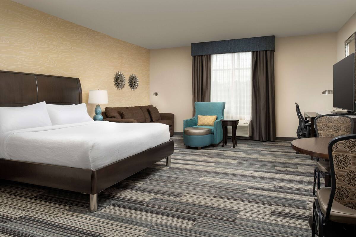 A hotel room with a large bed, a sofa, a chair, a small table, and a TV.