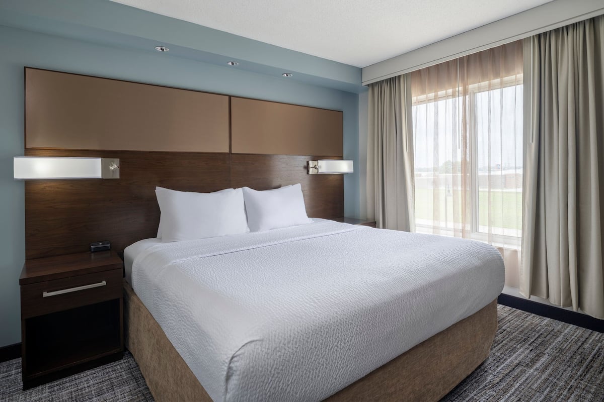 A hotel room with a large bed, a wooden headboard, two bedside tables, and a view of the outdoors through large windows.