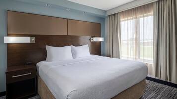 A hotel room with a large bed, a wooden headboard, two bedside tables, and a view of the outdoors through large windows.
