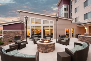 Un patio con mobiliario exterior, una fogata y un edificio con ventanas amplias y un muro de piedra.