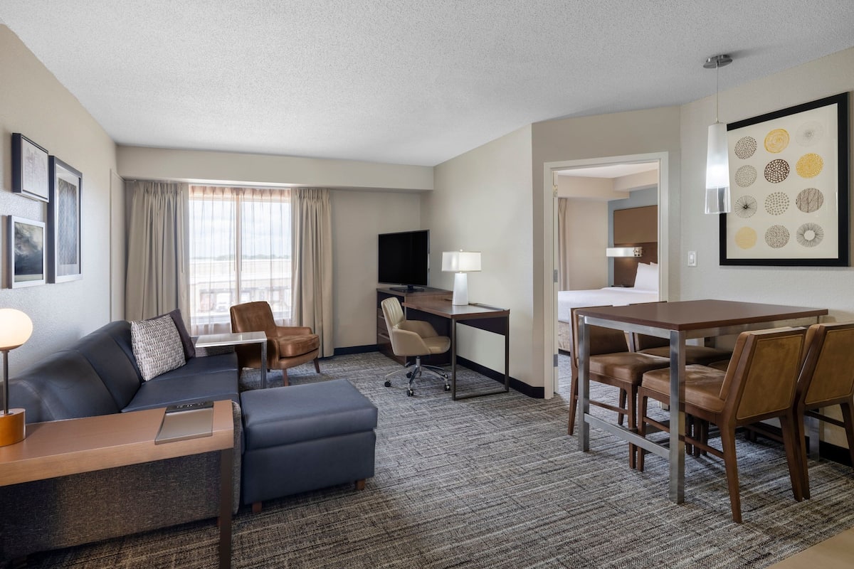 A hotel room with a sofa, a desk, a dining table with chairs, a TV, and a large window with curtains.