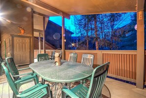 Een overdekt terras buiten met een ronde metalen tafel en groene stoelen, een houten deur, en een uitzicht op bomen en een huis op de achtergrond.
