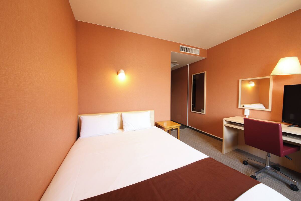 A hotel room with a bed, a desk, a chair, a mirror, and a wall-mounted lamp.