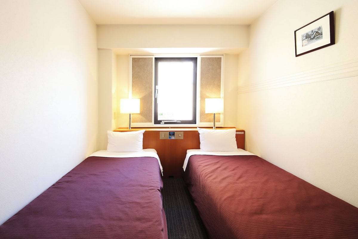 A hotel room with two single beds, a window with a view, and a small picture on the wall.