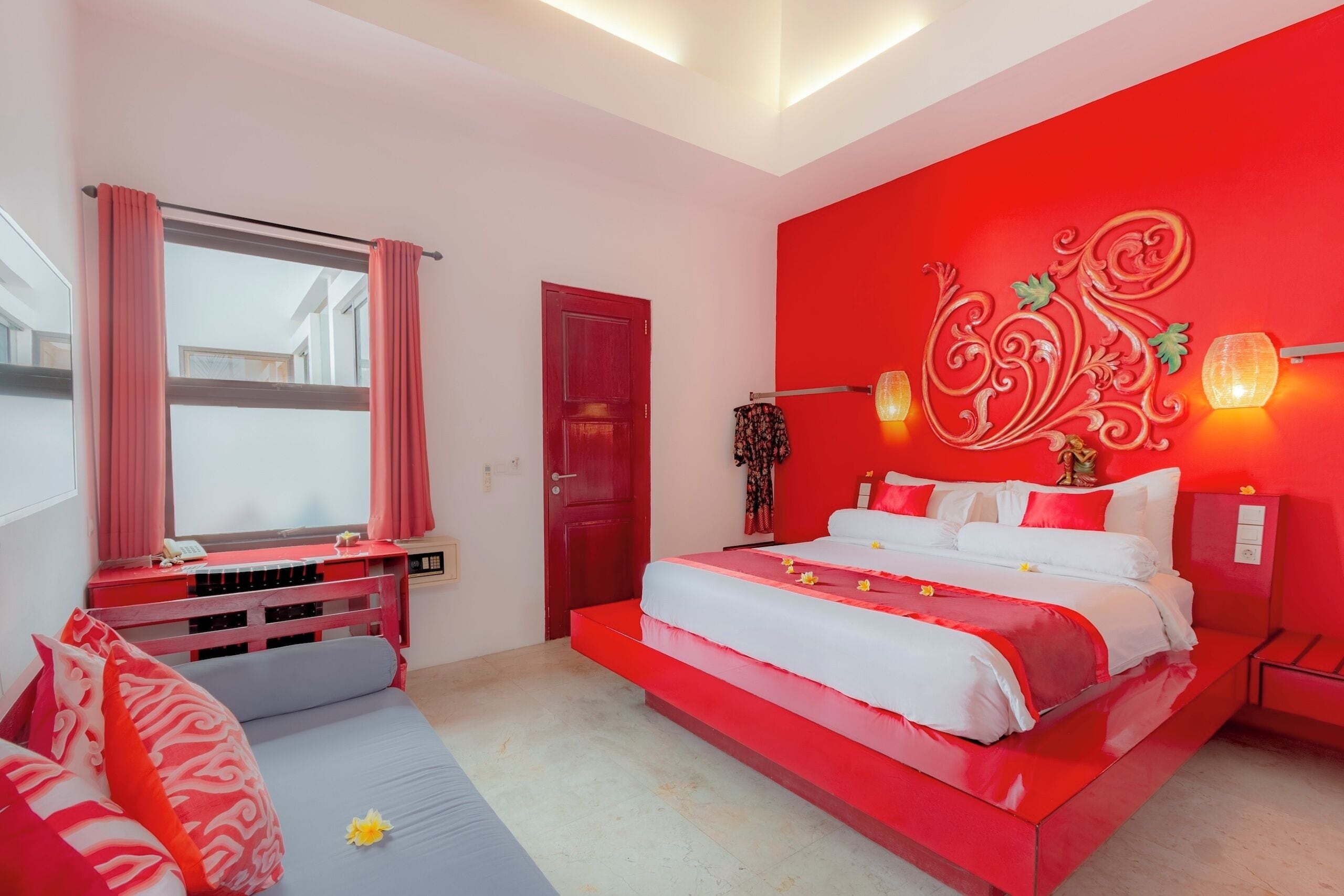 A bedroom with a red accent wall, a bed with white and red bedding, a desk with a computer, and a window with pink curtains.