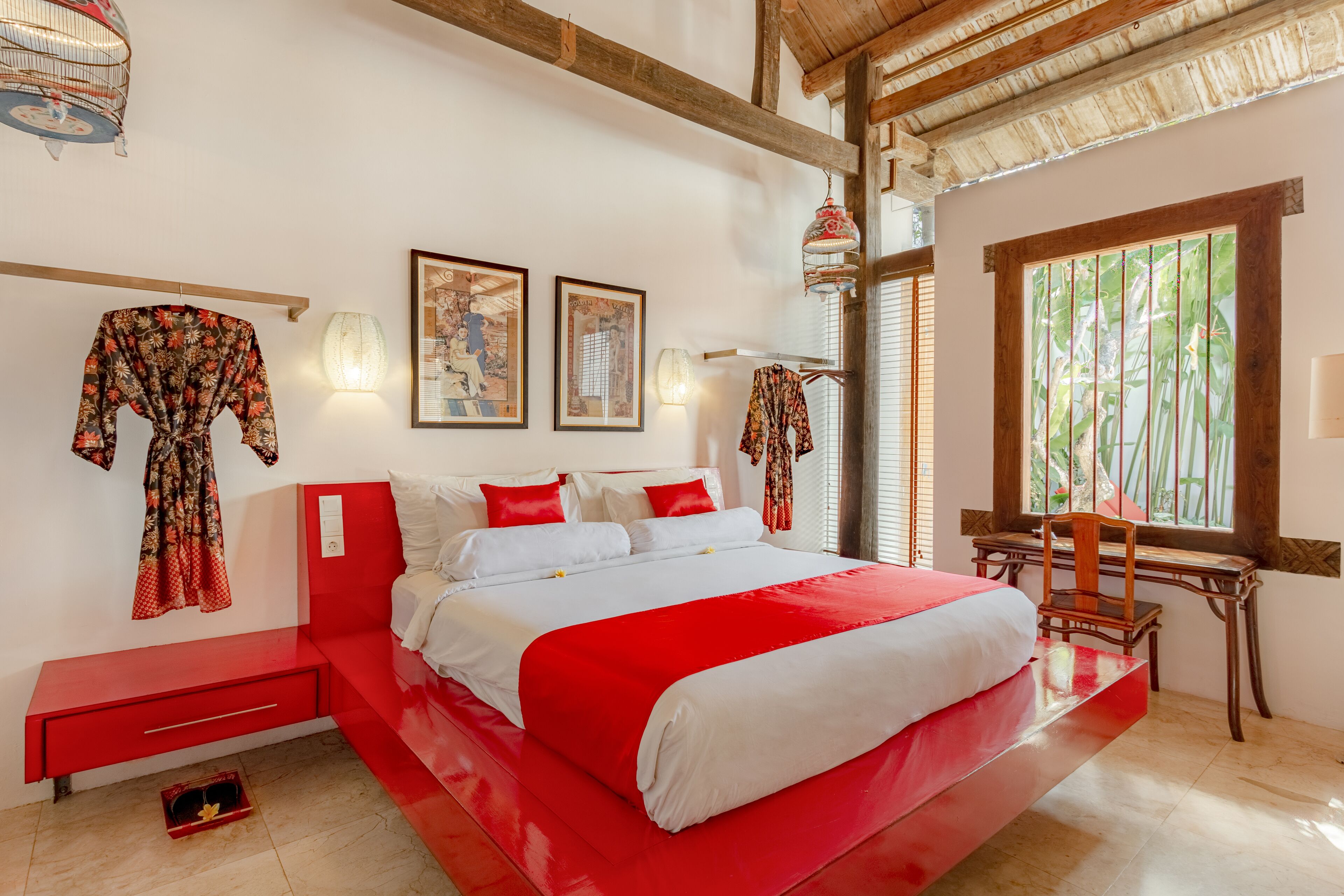 Un dormitorio con una cama grande, ropa de cama roja y blanca, un escritorio de madera y una ventana con vistas a follaje.