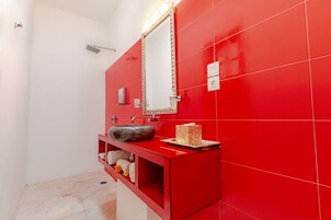 Casa de banho com parede revestida em azulejos vermelhos, um espelho grande, lavatório com cuba original e zona de duche.