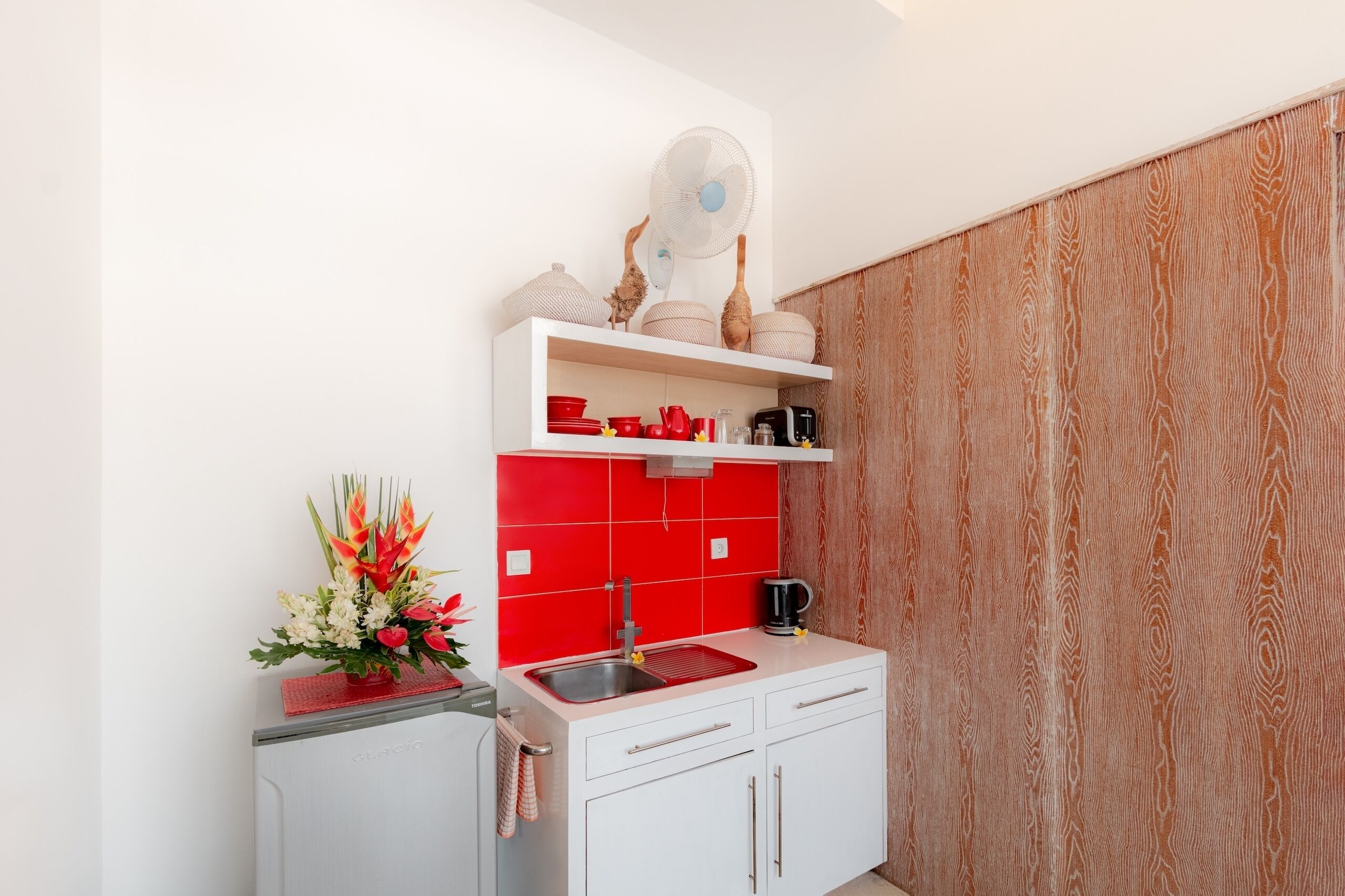 Una cocina compacta con salpicadero rojo, armarios blancos y panel de pared de madera.