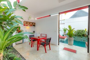 A modern kitchen with white cabinets, a red dining table, and chairs. There is a large potted plant on the left and a small table with a red bench near the glass door leading to a pool area. - Bali Ginger Suites & Villa (Seminyak)