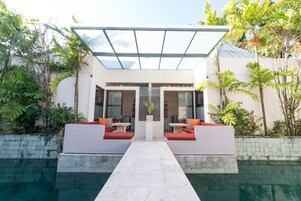Uma área moderna de piscina externa com teto de vidro, duas áreas de estar com almofadas vermelhas e plantas tropicais exuberantes.
