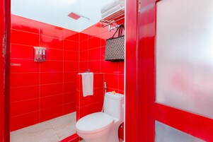 Casa de banho com azulejos vermelhos, sanita branca, um porta-toalhas e um saco pendurado.