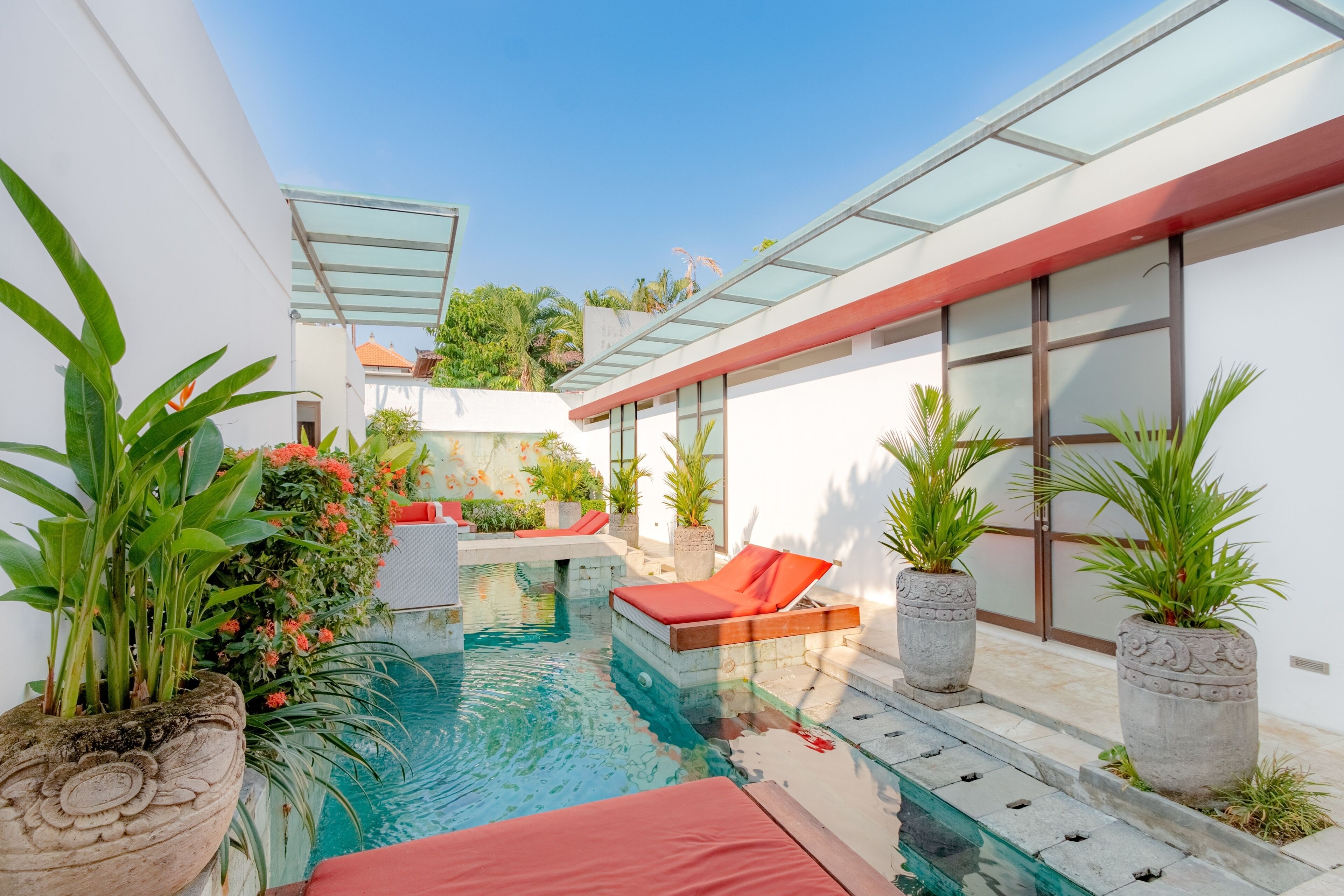 Un área de piscina exterior moderna con techo de vidrio, sillas reclinables rojas y plantas en macetas.