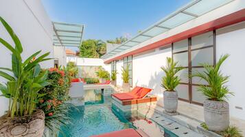 Un área de piscina exterior moderna con techo de vidrio, sillas reclinables rojas y plantas en macetas.