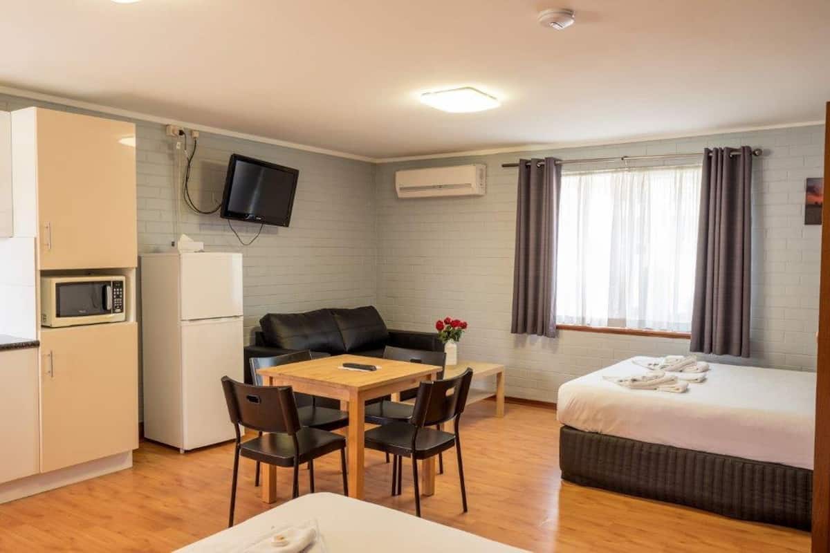 A compact hotel room with a kitchenette, a bed, a sofa, a dining table with chairs, and a TV mounted on the wall.