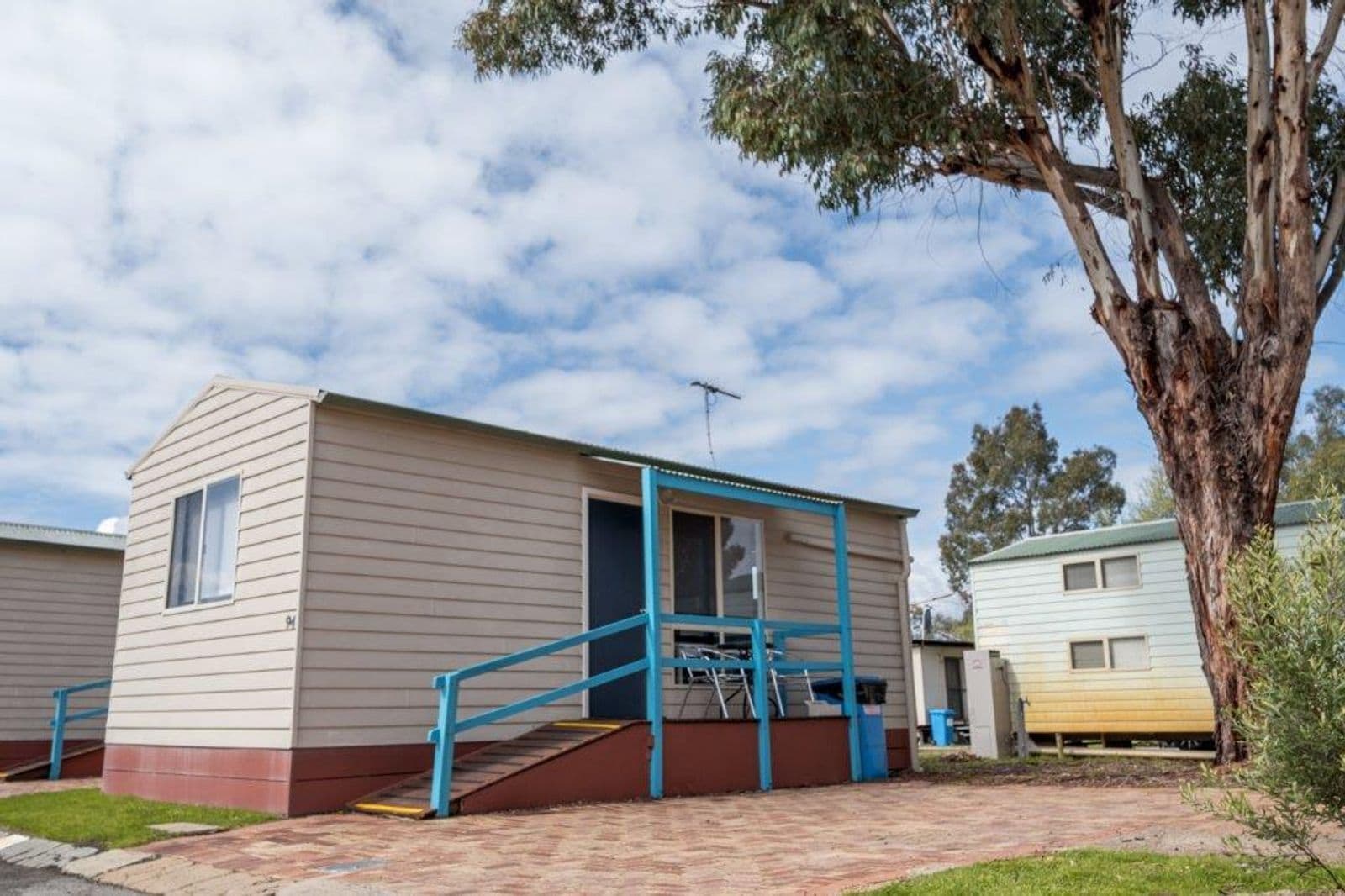 Une maison mobile de plain-pied, avec un escalier bleu menant à l’entrée, un petit porche et un arbre en arrière-plan.