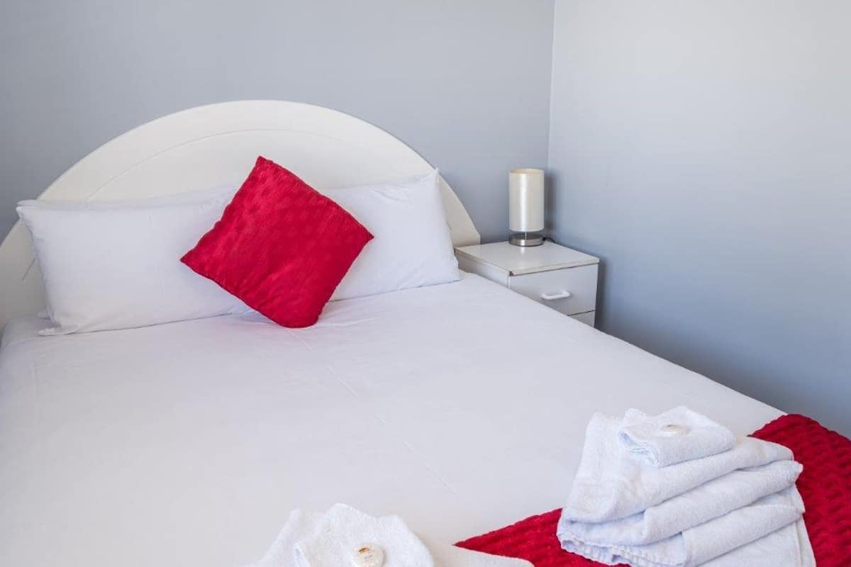 A neatly made bed with white bedding and a red pillow, a white bedside table with a lamp, and a red throw blanket on the floor.