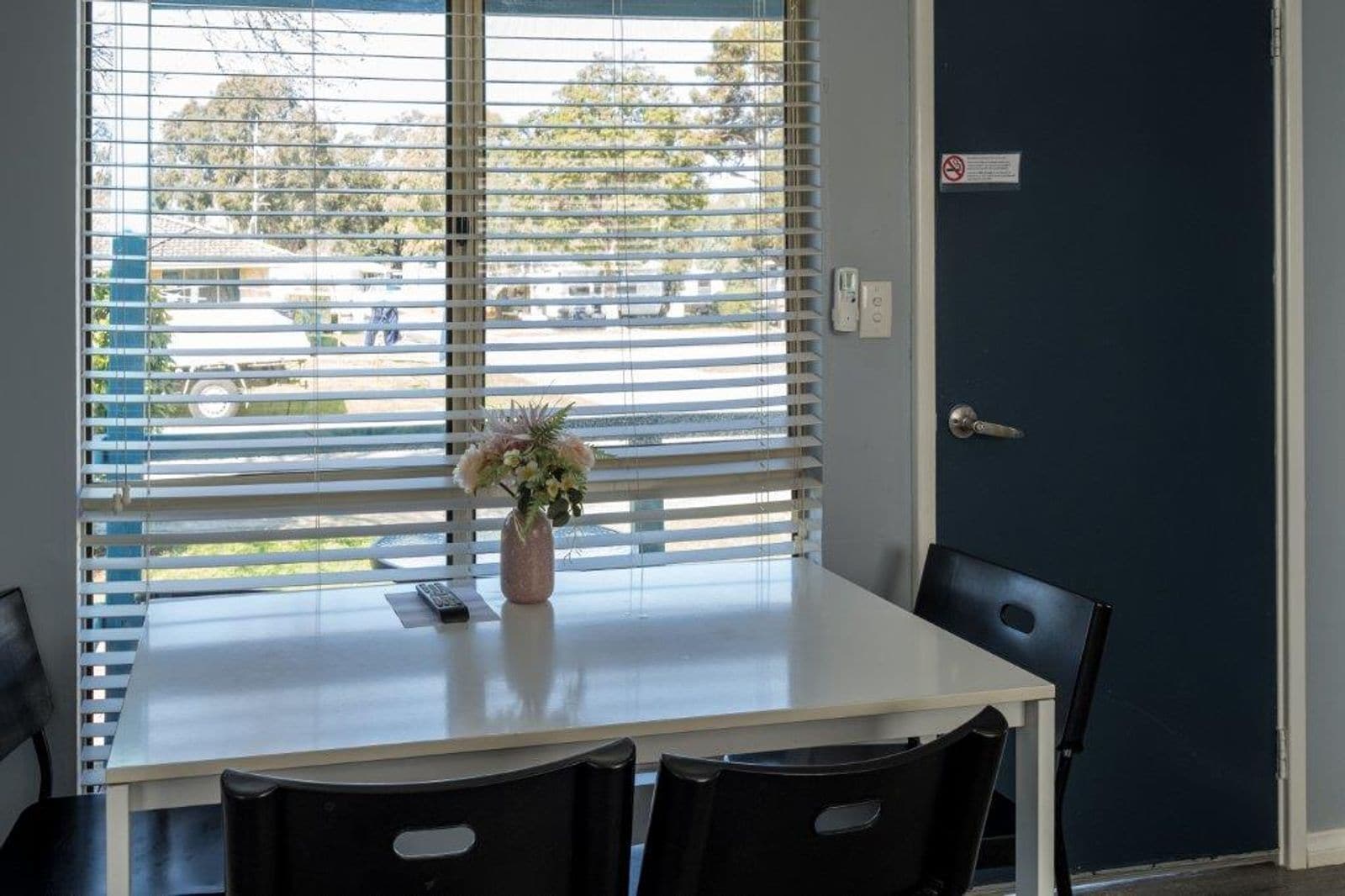 A dining area with a table, chairs, a vase with flowers, and a view through window blinds.