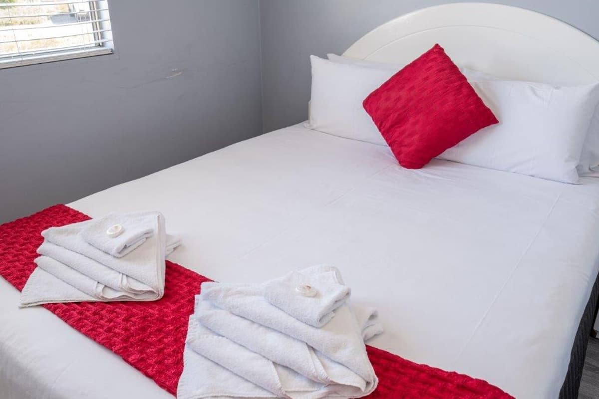 A single bed with white bedding and a red pillow, two neatly folded towels on a red mat, and a window with blinds.