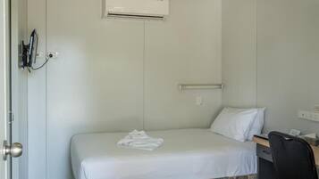 Un lit simple avec un matelas et des oreillers blancs, un petit bureau avec une chaise et un appareil de climatisation fixé au mur.