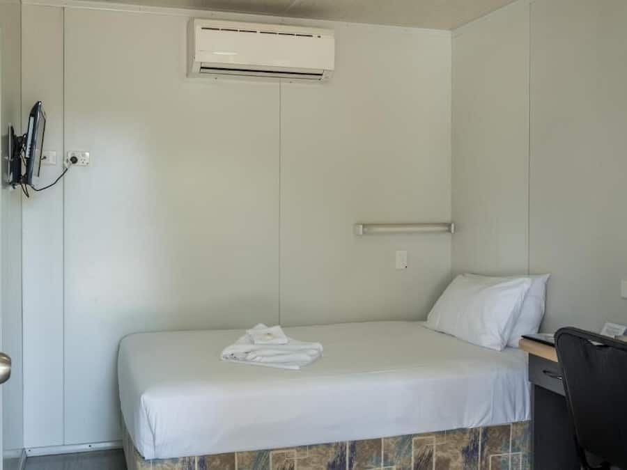 Una cama individual con colchón y almohadas blancos, un escritorio pequeño con una silla, y un equipo de aire acondicionado instalado en la pared.