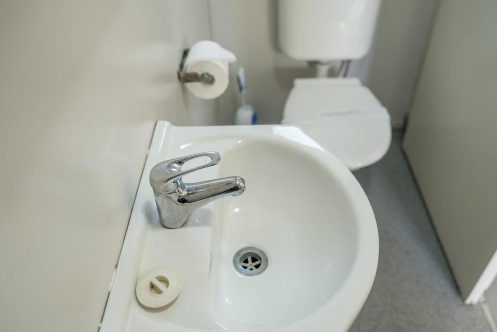 A white bathroom sink with a chrome faucet, a toilet paper holder, and a toilet in the background.