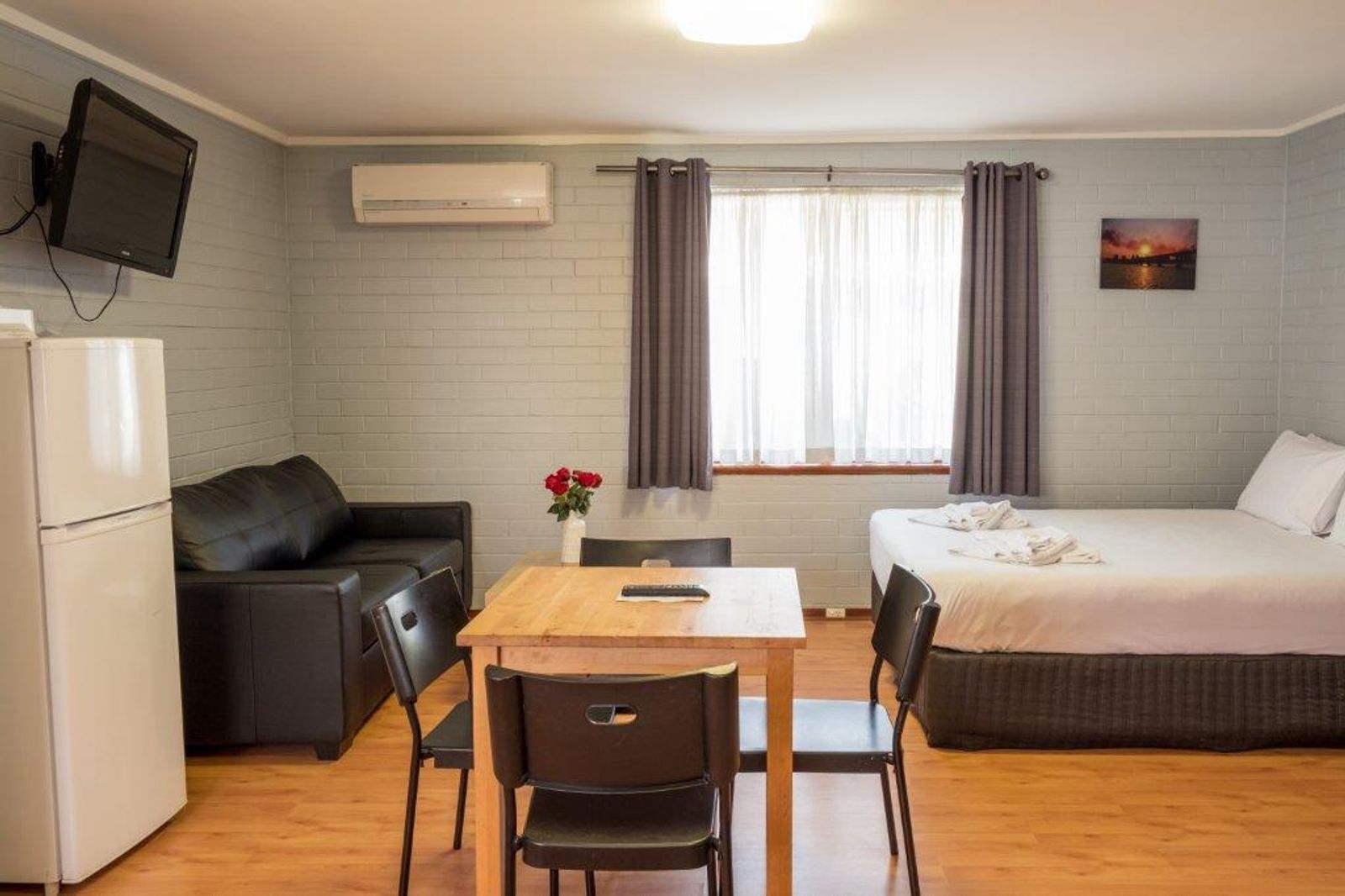 Ein kompaktes Hotelzimmer mit Bett, Sofa, Esstisch mit Stühlen, einem an der Wand befestigten Fernseher und einer Klimaanlage.