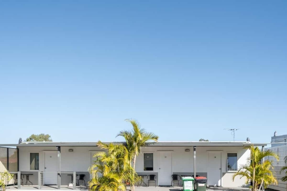 A row of single-story motel rooms with individual door numbers, outdoor seating, and a clear blue sky.