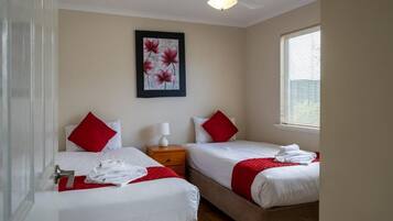 A hotel room with two beds, each with red pillows and white bedding. There is a ceiling fan, a framed artwork on the wall, and a window with blinds.