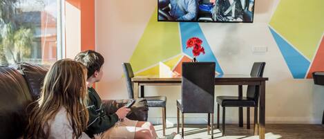 A room with a large TV mounted on the wall, a couch, a dining table with chairs, and a vase with flowers on the table.