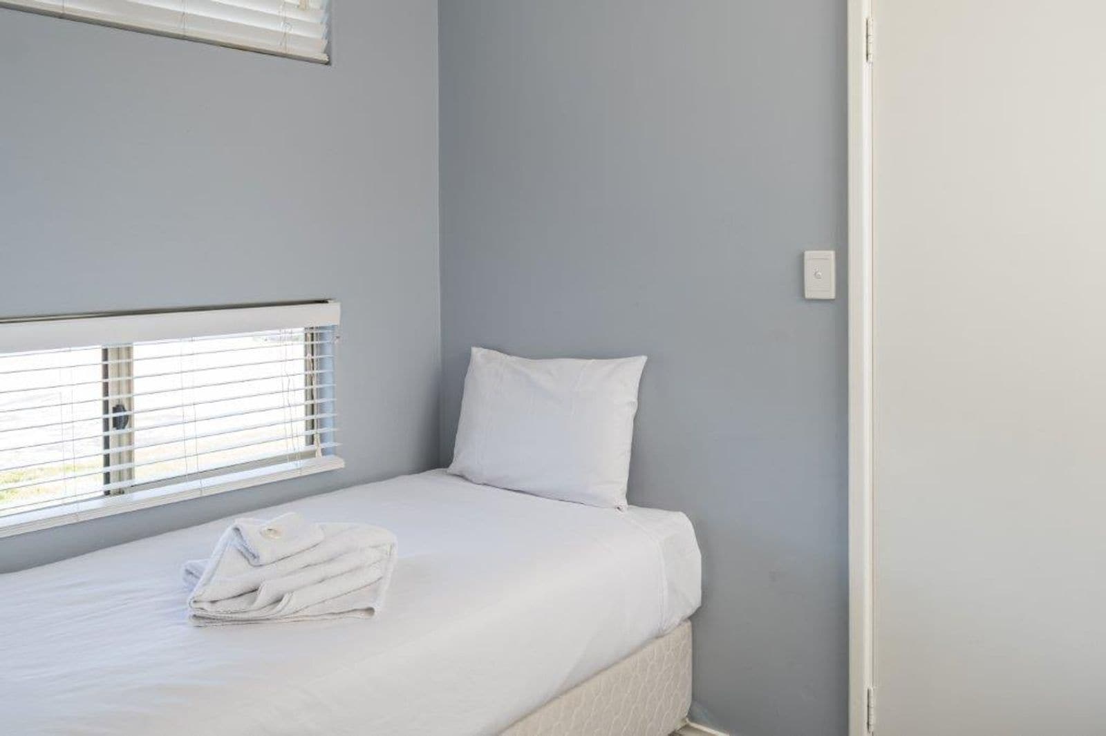 Una cama individual con ropa de cama blanca y una toalla doblada, una ventana con persianas y una pared lisa.