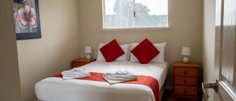 A neatly made bed with white and red bedding, two bedside tables with lamps, and a window with blinds.