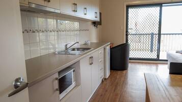 Una cocina compacta con armarios blancos, un lavamanos de acero inoxidable y un microondas. Hay una mesa de madera y un bote de basura negro.