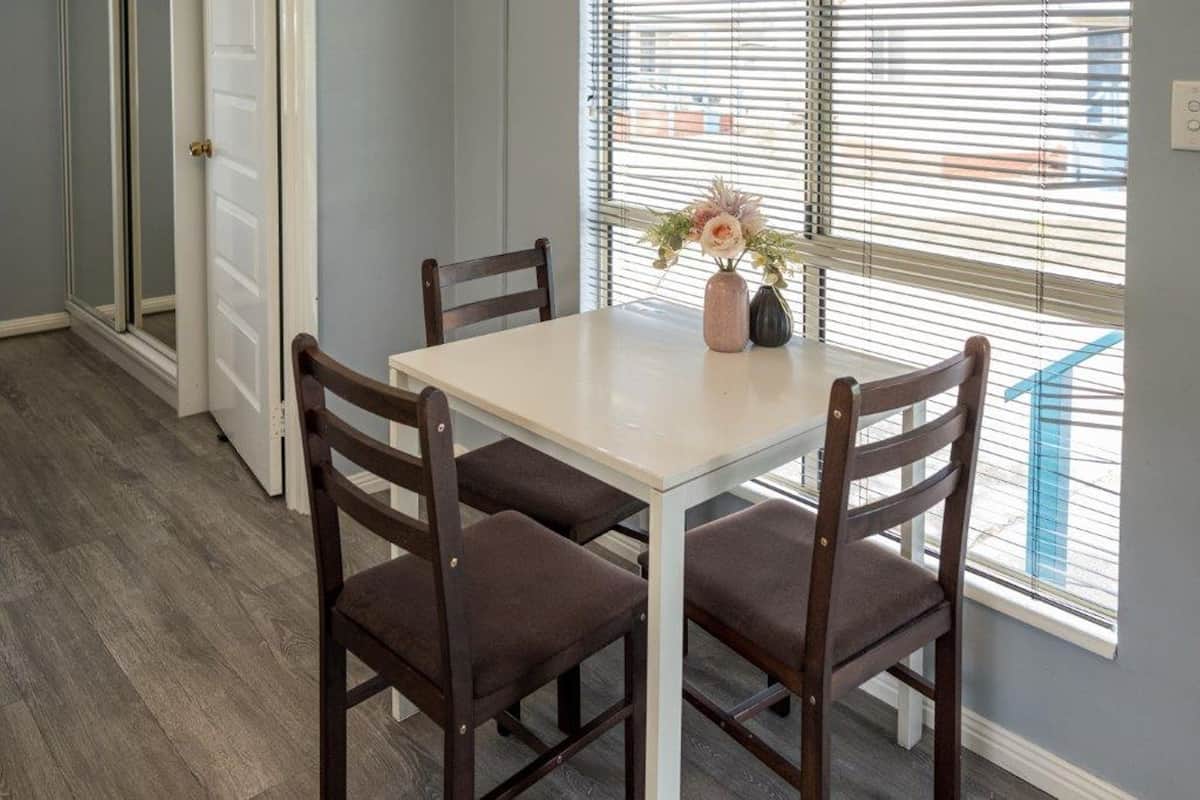 A dining area with a table and four chairs, a vase with flowers, and a window with blinds.