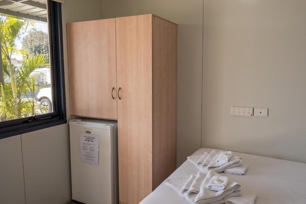 A small room with a wooden wardrobe, a white refrigerator, and a bed with neatly folded white towels.