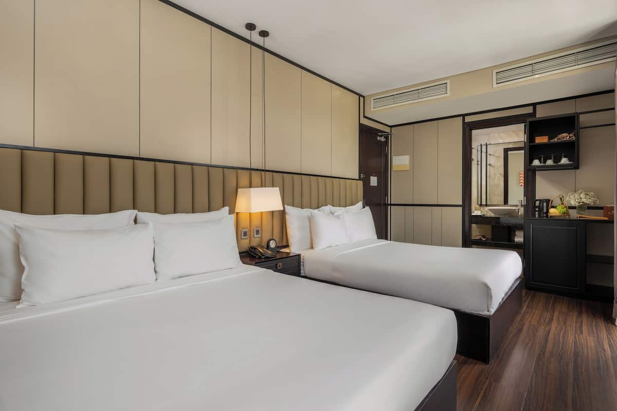 A hotel room with two beds, a nightstand, a lamp, a mirror, and a small kitchenette.
