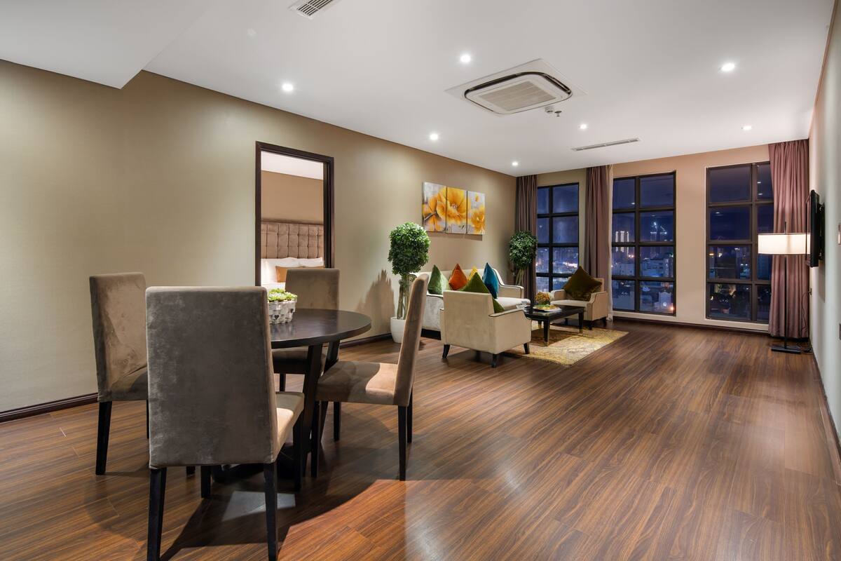 A modern hotel room with a dining area, a large window with a city view, and a flat-screen TV mounted on the wall.