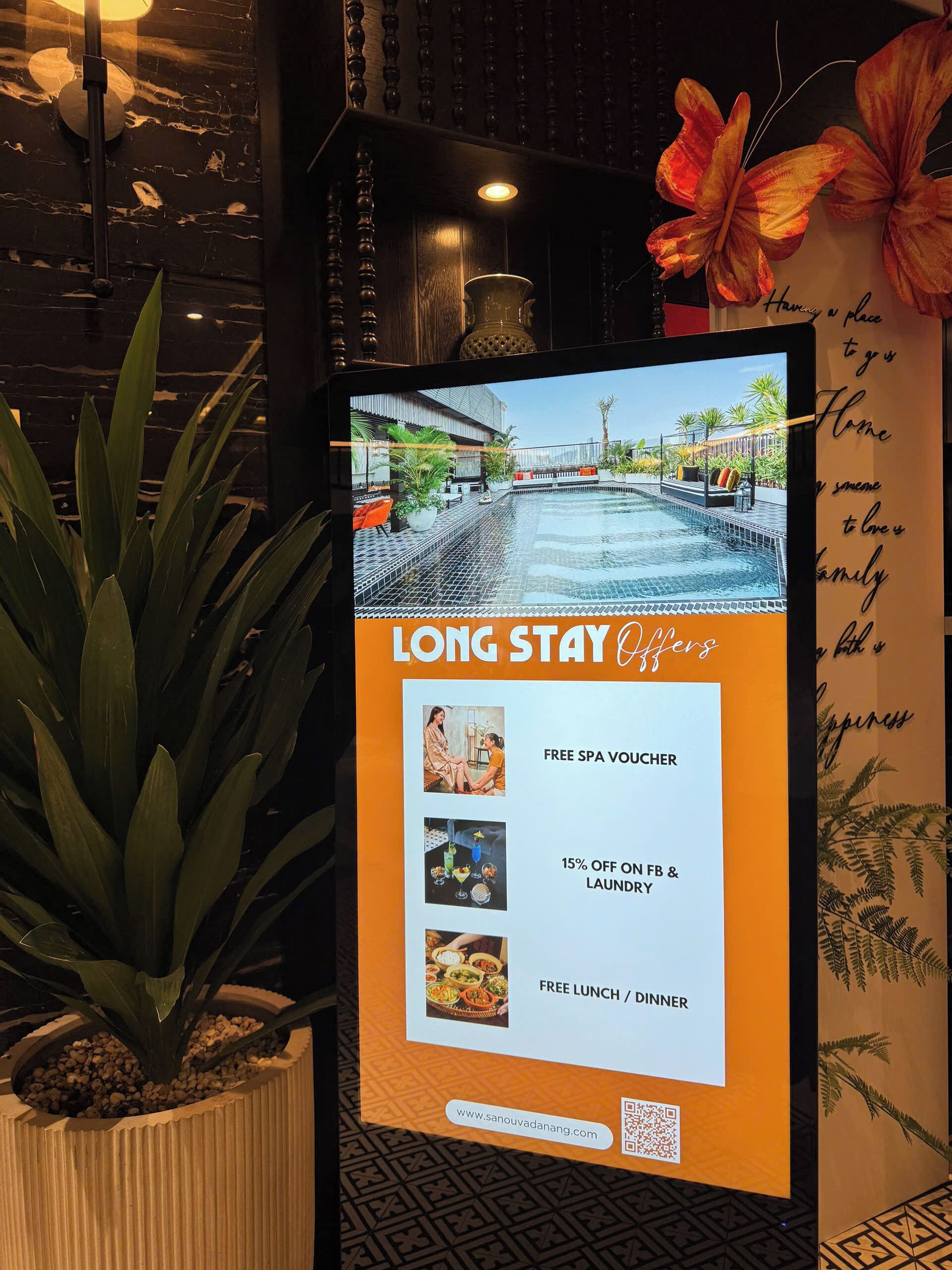 A digital display board advertising a long stay offer with amenities like a free spa voucher, 15% off on food and laundry, and free lunch/dinner.