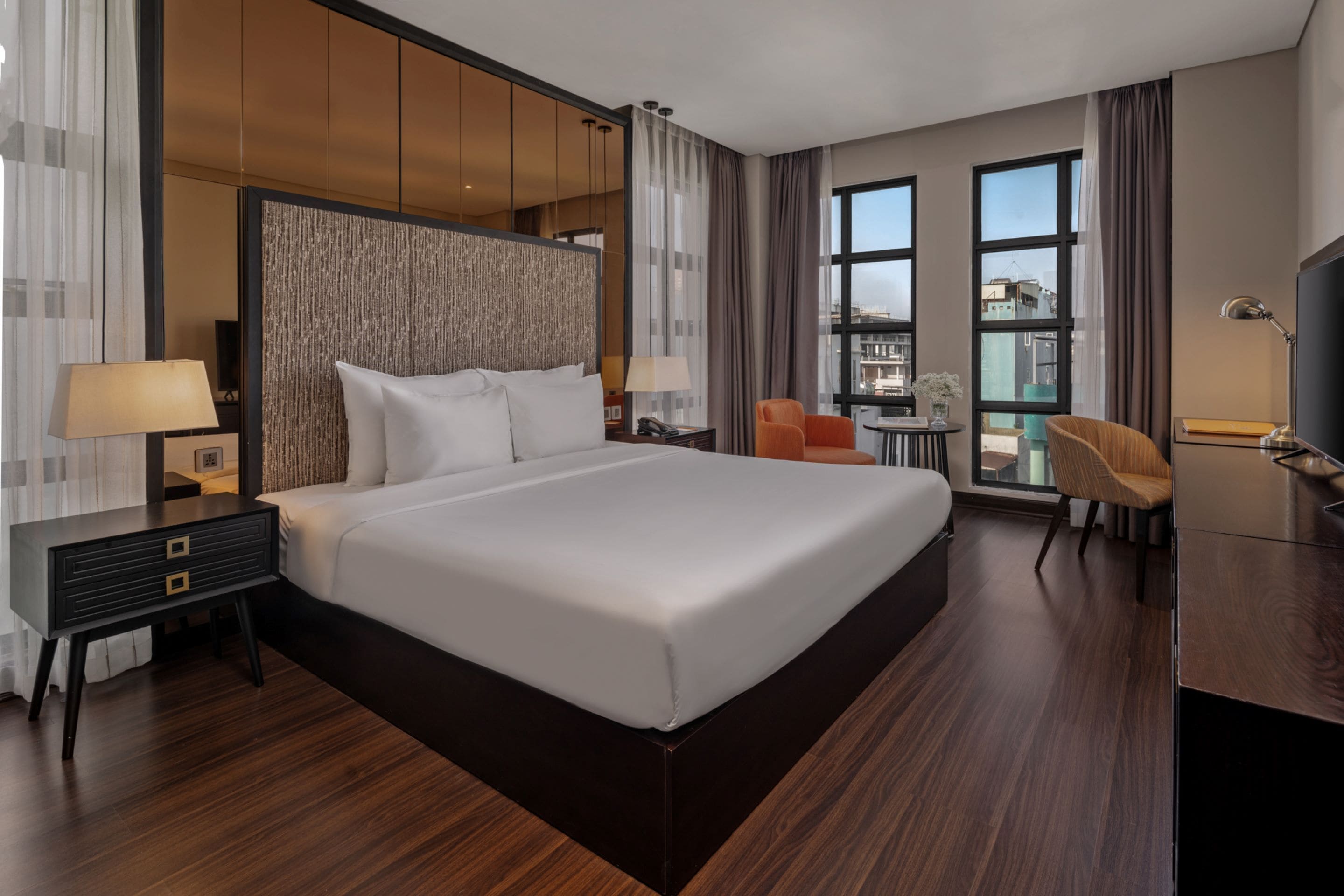 A modern hotel room with a large bed, a desk with a lamp, a chair, and a TV.