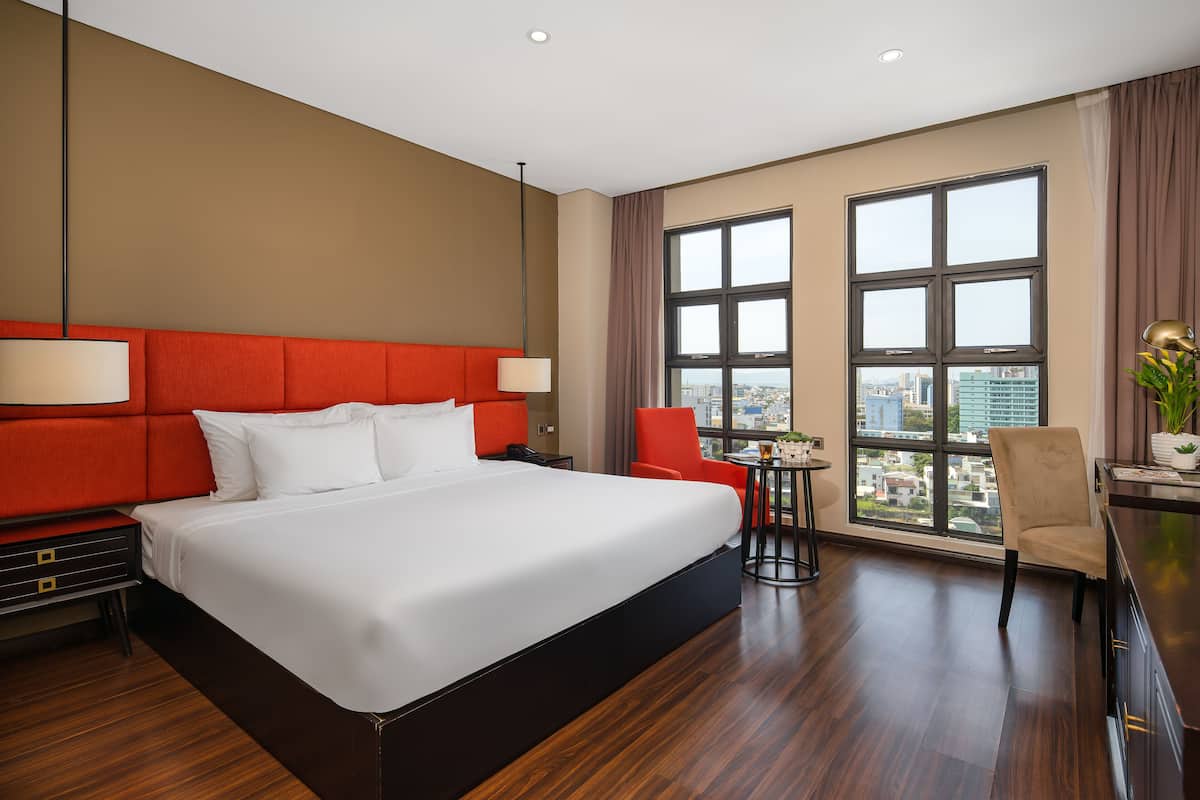 A hotel room with a large bed, red headboard, a desk with a chair, and a view of the city through large windows.