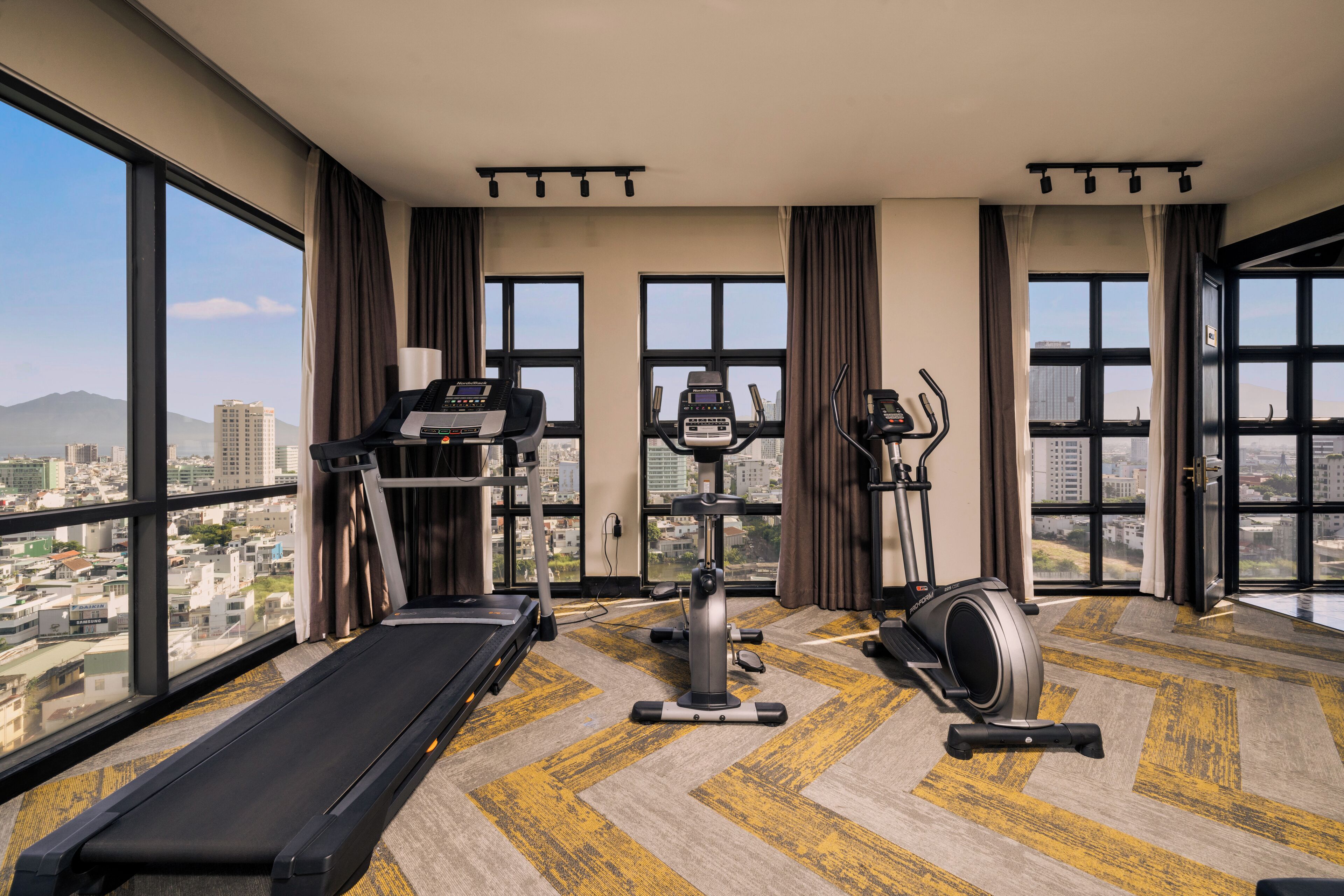 A gym with treadmills, elliptical machines, and stationary bikes, large windows with a city view.
