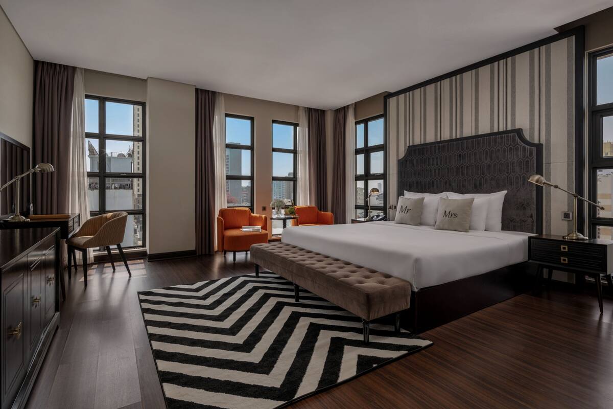 A modern hotel room with a large bed, two chairs, a desk, and a view of the city through large windows.