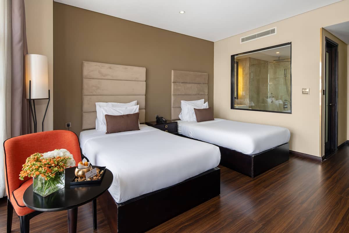 A hotel room with two beds, a small table with a vase of flowers, a chair, and a large mirror.