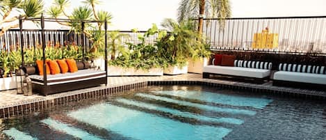 Un área de piscina en la azotea con un conjunto de sofá blanco y negro, una zona de estar pequeña con cojines anaranjados y diversas plantas en macetas.
