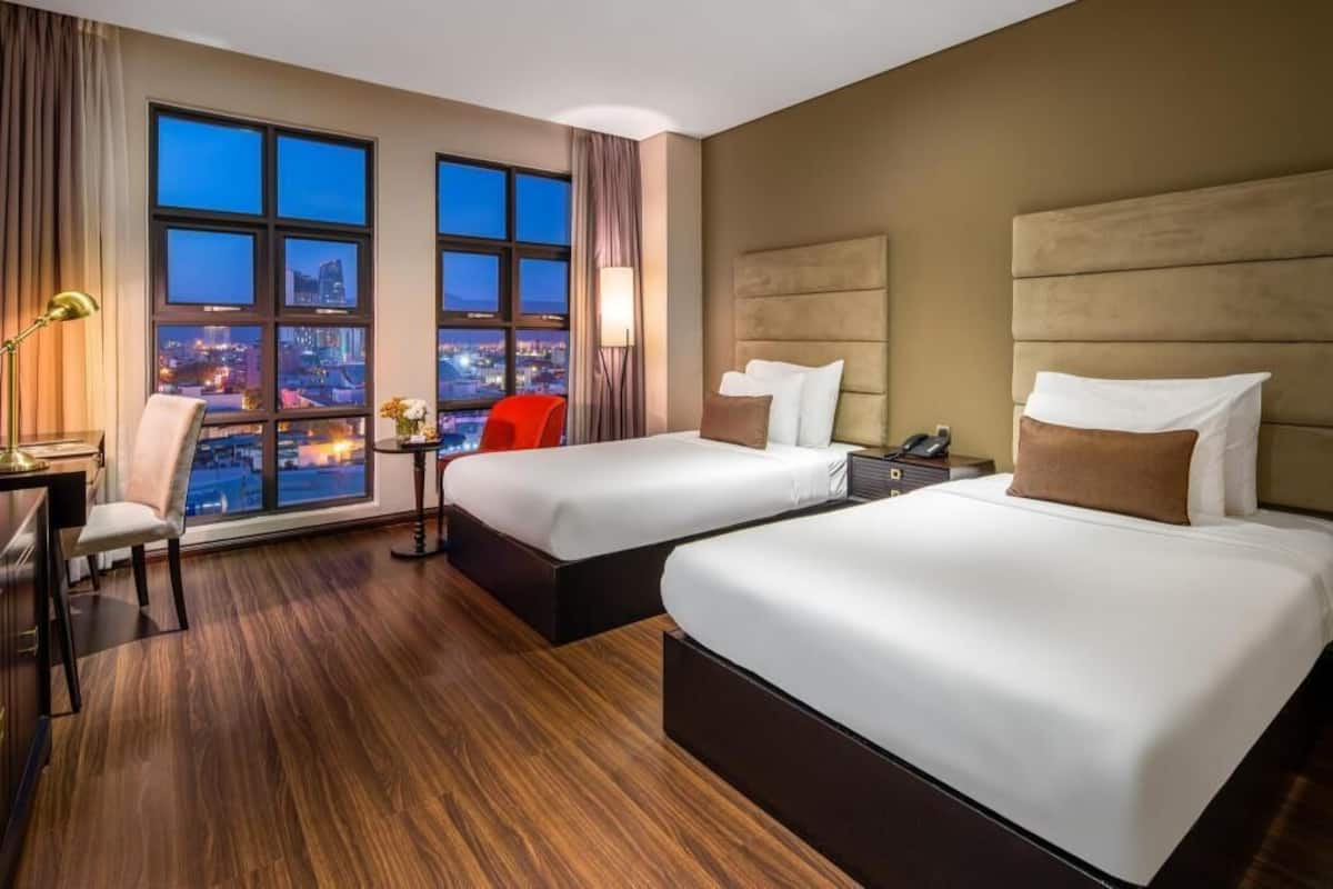 A hotel room with two beds, a desk, a chair, a nightstand, and a large window with a city view.